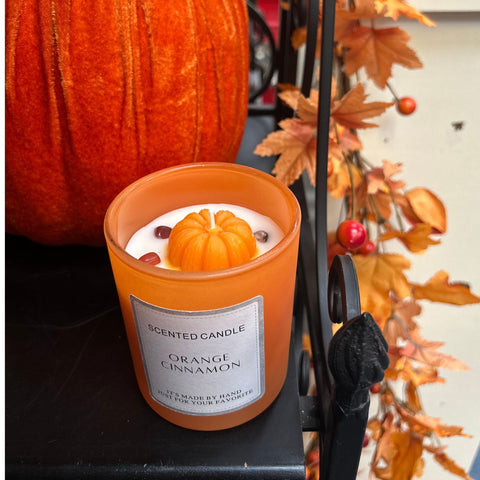 Orange cinnamon scented candle with a pumpkin design on top, surrounded by autumn leaves and a pumpkin.