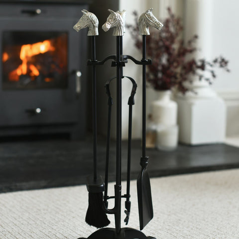 Set of fireplace tools with horse heads on a stand in front of a lit fireplace.