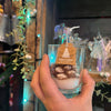Hand holding a small glass with a candle and decorative tag against a festive background.