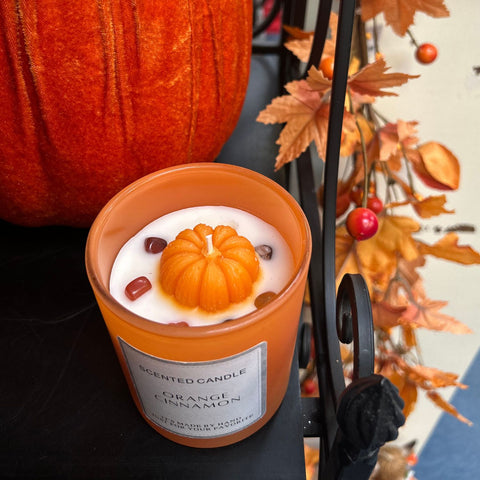 Scented candle with pumpkin design in front of a pumpkin and autumn leaves.