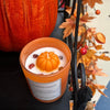 Scented candle with pumpkin design in front of a pumpkin and autumn leaves.