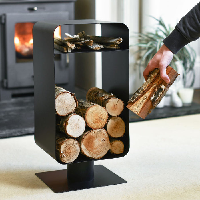 Black firewood holder with logs and a person placing more wood inside, with a fireplace in the background.