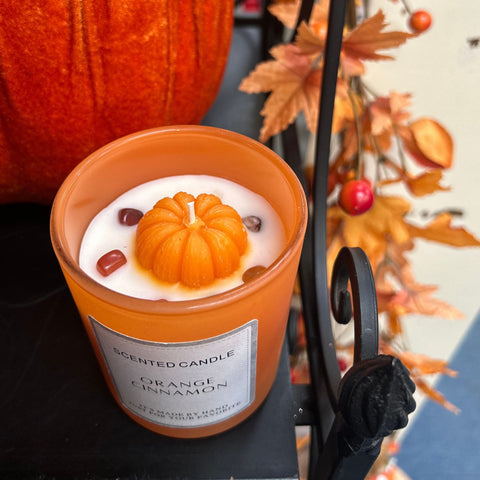 Scented candle with pumpkin design in an orange container, surrounded by autumn leaves and berries.