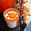 Scented candle with pumpkin design in an orange container, surrounded by autumn leaves and berries.