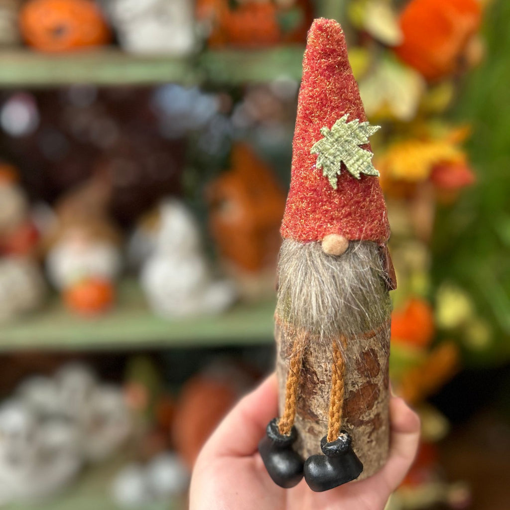 Hand holding a small gnome figurine with a red hat and autumn-themed decorations in the background.