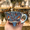 Ceramic teapot with bird and berry design held in a hand, with a blurred store background.