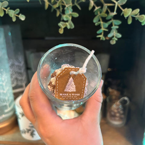 Hand holding a small glass container with a candle inside, featuring a 'Make a Wish' tag.