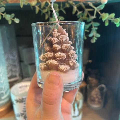 Hand holding a small glass candle with a pine cone design against a blurred indoor background