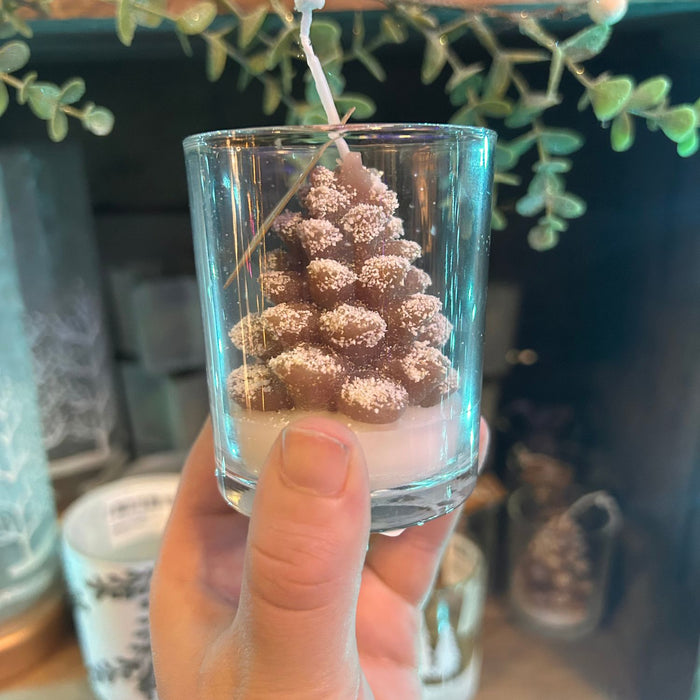 Hand holding a small glass candle with a pine cone design against a blurred indoor background
